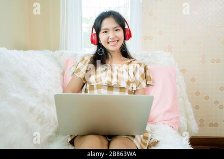 Asian girls are listening to music, Red headphones. And she is using a laptop to work, Sitting on a white sofa in her room. Stock Photo