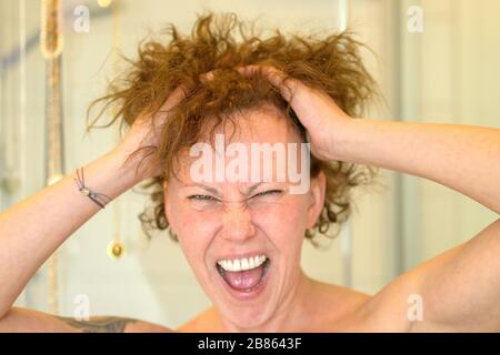 Frustrated woman having a bad hair day running her hands through her unruly curly hair and yelling in a close up head shot Stock Photo