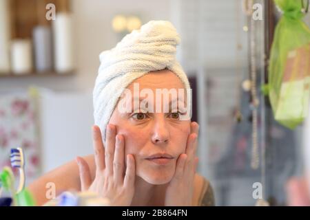Woman wearing a towel on her wet hair after showering taking a close look at her face in a bathroom mirror in a personal hygiene concept Stock Photo
