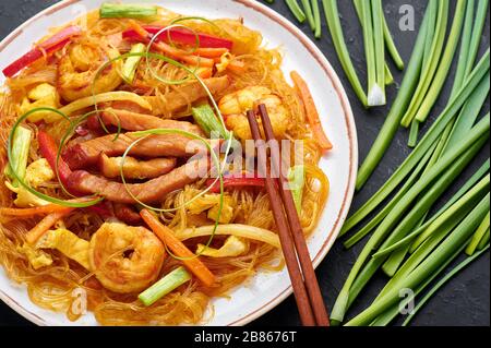 Singapore Mei Fun in white plate on dark slate background. Singapore Noodles is chinese cuisine dish with rice noodles, prawns, char siu pork, carrot, Stock Photo