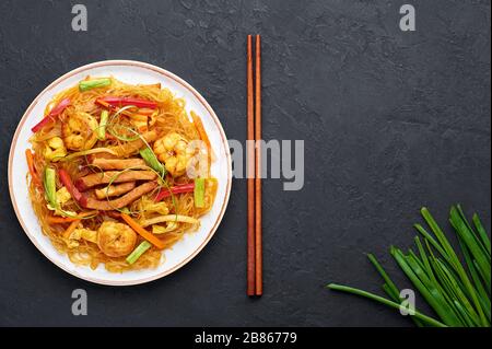 Singapore Mei Fun in white plate on dark slate background. Singapore Noodles is chinese cuisine dish with rice noodles, prawns, char siu pork, carrot, Stock Photo