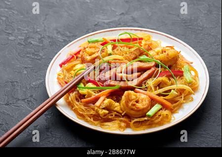 Singapore Mei Fun in white plate on dark slate background. Singapore Noodles is chinese cuisine dish with rice noodles, prawns, char siu pork, carrot, Stock Photo