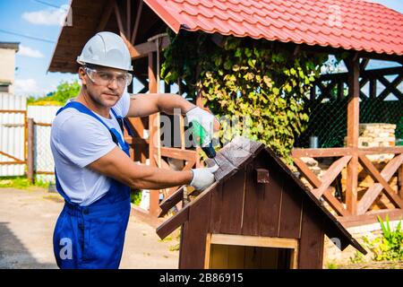 Carpenter using electric screwdriver. Man builds a roof of wooden planks of a small house. Building dog booth. Countryside house. Stock Photo