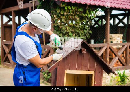 Carpenter using electric screwdriver. Man builds a roof of wooden planks of a small house. Building dog booth. Countryside house. Stock Photo