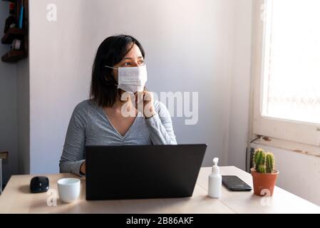 young woman wearing protective face mask and with hand sanitizer in home office looking worried and stressed out of the window due to corona virus Stock Photo