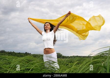 Young smiling curly woman in white clothes standing with closed eyes and holding yellow silk shawl in raised hands in meadow and green grass in summer Stock Photo