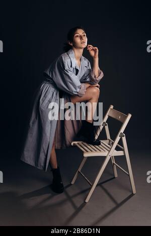 Brunette girl in trench and butylons rests with one foot on a chair Stock Photo