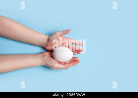 White chicken egg in the hands of a child on a blue background. Easter concept. Stock Photo