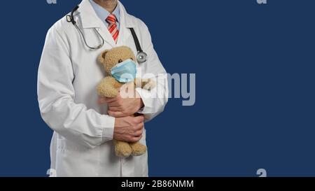 Medical doctor with a stethoscope on shoulders holds a teddy bear in a medical mask against blue background with pace rof text Stock Photo
