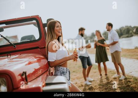 Happy young women drinks cider from the bottle by the convertible car with friends Stock Photo