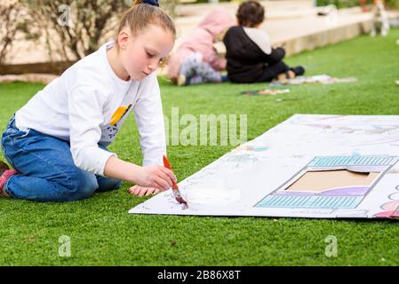 Kids play outdoors. Child girl paints a paper house template with colorful watercolors on the grass in the garden. Stock Photo