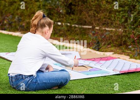 Stress relief with art therapy. Back view teenager girl paints a paper house template with colorful watercolors on the grass in the garden. Homeschool, child play outdoor play-area on tne backyard. Stock Photo