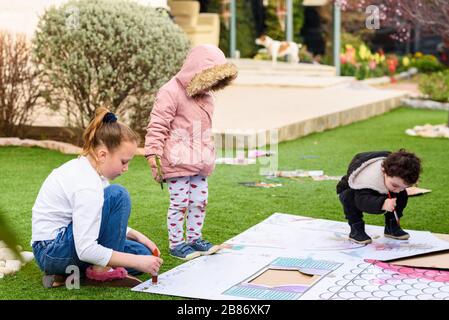 Kids play outdoors. Children paints a paper house template with colorful watercolors on the grass in the garden. Stock Photo