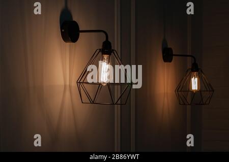 Pendant lights made of diamond shape wire carcass with glass bulb inside. Glowing filament in lamps of minimal style. Sconce lamp hanging on wall. Stock Photo