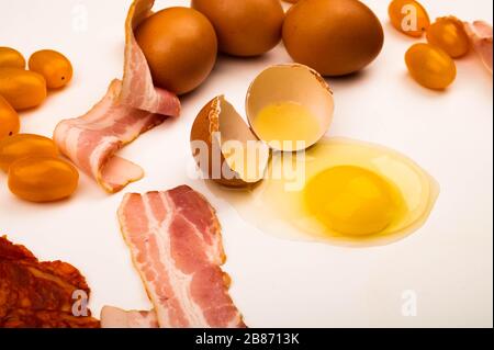 Broken chicken egg, scattered chicken eggs, tomatoes and slices of sausage and bacon on a white background. Close up Stock Photo