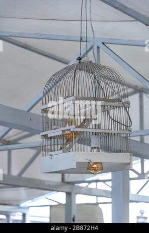 Lampshade made from white painted wooden bird cage. Closeup of birdcage transformed into ceiling lamp with golden color glass lightbulb inside. Stock Photo