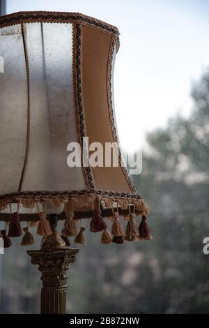 Matte transparent vintage lampshade with tassels by bottom edge in front of blurred view through window. Old-fashioned antique table lamp closeup. Stock Photo