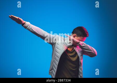 Winner's gesture, dab, dabbing. Caucasian man's portrait isolated on blue studio background in pink neon light. Beautiful male model in casual. Concept of human emotions, facial expression, sales, ad. Stock Photo