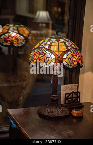 Antique table lamp with stained glass lampshade closeup on dark blurry background. Glowing mosaic lamp in shape of dome. Vintage table-lamp. Stock Photo