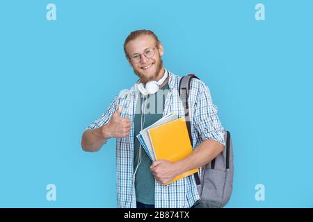 Happy male student hold backpack showing thumbs up isolated on blue background. Stock Photo