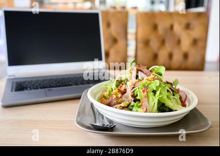 Modern laptop with spicy pork salad on wooden table Stock Photo