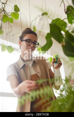 Young woman in eyeglasses holding bottle and watering flowers in her room Stock Photo