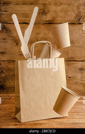 Flying disposable paper tableware over bag with mock up on wooden background. Environmental care concept. Stock Photo