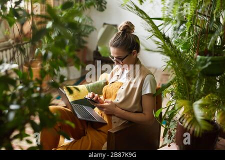 Young florist in eyeglasses sitting on armchair with laptop computer on her knees and using mobile phone in the room with plants Stock Photo