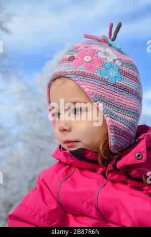 Portrait of young girl in ski sportswear and colorful woolen hat, looking away, against blue sky and white, frozen trees bokeh Stock Photo