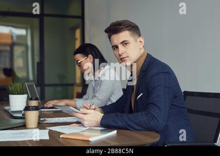 Portrait of young businessman looking at camera and working with tablet pc at the table with businesswoman in the background at office Stock Photo