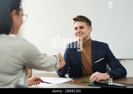 Young smiling businessman sitting at the table and shaking hand to young businesswoman during the meeting at office Stock Photo
