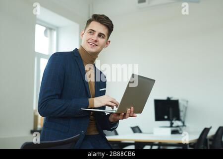 Young smiling businessman standing and holding laptop computer in his hands he working online at office Stock Photo