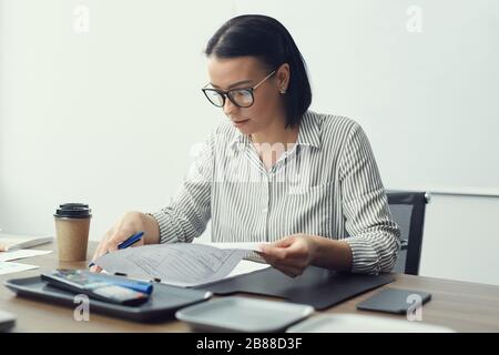 Young businesswoman in eyeglasses sitting at the table with coffee cup and doing paperwork at office Stock Photo