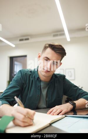 Young man sitting at the table and making notes in notepad while working at office Stock Photo