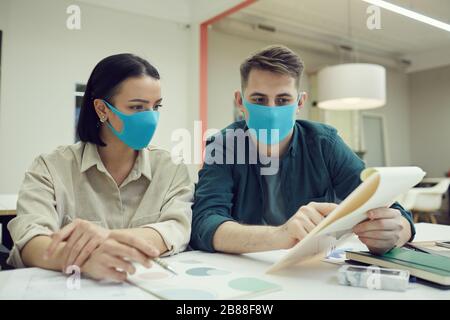 Two business colleagues wearing protective masks and discussing business document together at the table at office Stock Photo