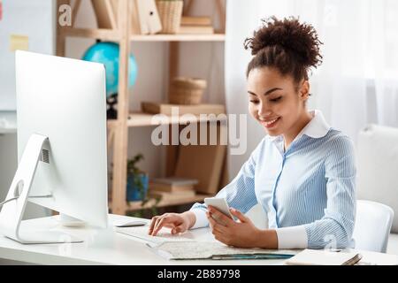 Tour agent sitting at table in office browsing computer looking at new message on smartphone happy Stock Photo