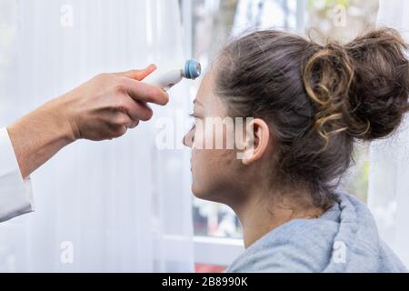Doctor in white medical coat measuring temperature from young woman using forehead digital thermometer in hospital room by window with bright natural Stock Photo