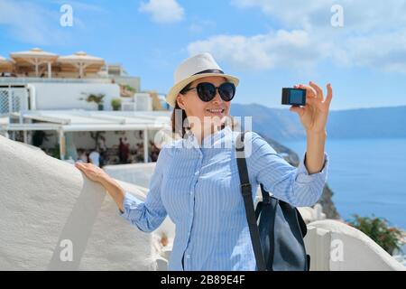 Woman travel vloger traveling in Greek village of Oia on Santorini island, filming aktion camera video, background white architecture, sea, sky in clo Stock Photo