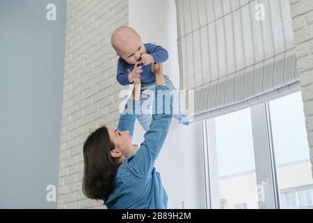 Happy young beautiful mother playing with toddler son, woman raising baby up, flying and laughing boy. Family is happy at home, near window, copy spac Stock Photo