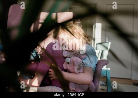 young girl curled up on a chair looking tired holding her toy at home