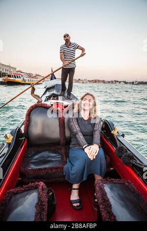 A woman posing for a picture on a gondola ride with the gondolier steering the boat in the background, Grand Canal, venice, Italy. Stock Photo