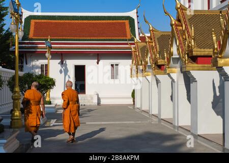 Two orange-robed Buddhist monks walking in the grounds of Wat Ratchanadta, the base of the iconic Lohaprasad building on the right; Bangkok, Thailand Stock Photo