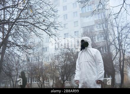 Side view of ecologist in protective uniform standing on foggy street and looking aside. Male environmentalist wearing white suit and gas mask. Concept of ecology and environmental pollution.