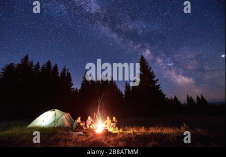Night camping near bright fire in spruce forest under starry magical sky with milky way. Group of four friends sitting together around campfire, enjoying fresh air near tent. Tourism, camping concept. Stock Photo