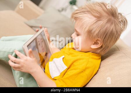 Close-up of cute son with blond hair sitting on sofa and using tablet while chatting with mom online Stock Photo