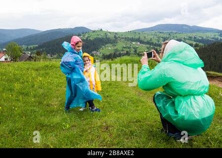 Mom in a raincoat takes pictures of her two pretty children in raincoats against the backdrop of picturesque meadows of hills and mountains in the cou Stock Photo