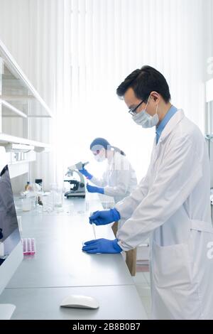 Young Chinese pharmacist or researcher testing new vaccine while his colleague studying virus characteristics on background Stock Photo
