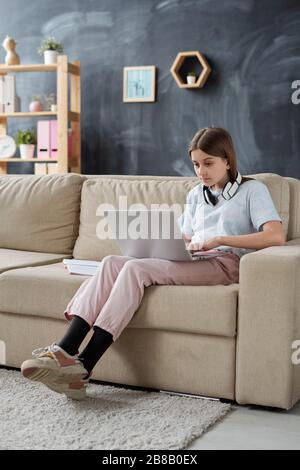 Cute female teenage student in casualwear looking at laptop display while relaxing on couch in living-room after school Stock Photo