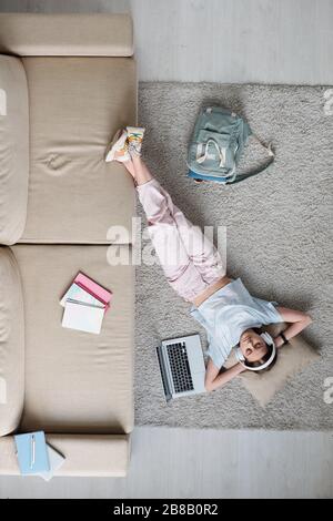 Overview of relaxed teenager in casualwear and headphones listening to music while relaxing on the floor by couch at home Stock Photo