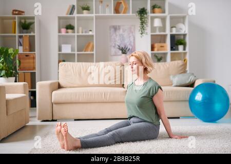 Mature blond active woman in sportswear sitting on the floor and exercising in living-room while staying at home for quarantine Stock Photo